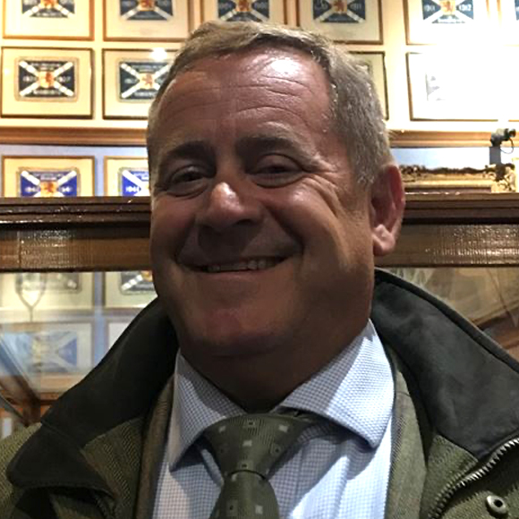 A smiling man dressed in formal attire, including a light blue shirt and patterned tie, standing indoors with a display of framed artwork in the background.