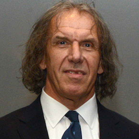 A professional headshot of a man with shoulder-length wavy hair, wearing a dark suit, white shirt, and a patterned tie, smiling slightly against a neutral background.