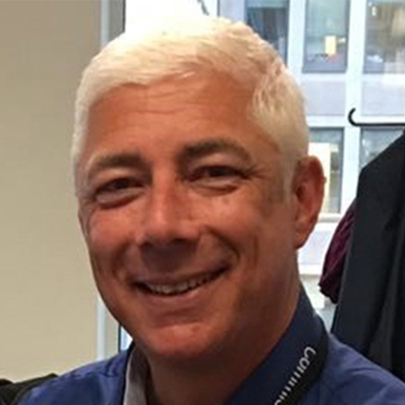 A smiling man with short white hair, wearing a blue collared shirt and a lanyard, with an office window in the background.