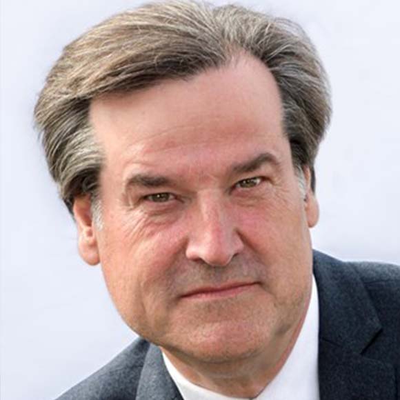 A portrait of a man with neatly styled gray hair, wearing a dark suit and white shirt, looking directly at the camera with a neutral expression.