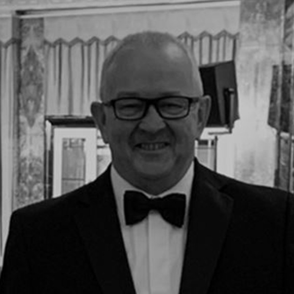 A man wearing glasses and a tuxedo with a bow tie, smiling, in a black-and-white photograph with an elegant interior background.