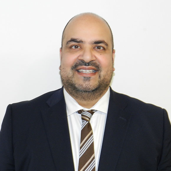 A bald man with a beard, wearing a dark suit, white shirt, and striped tie, smiling against a plain white background.