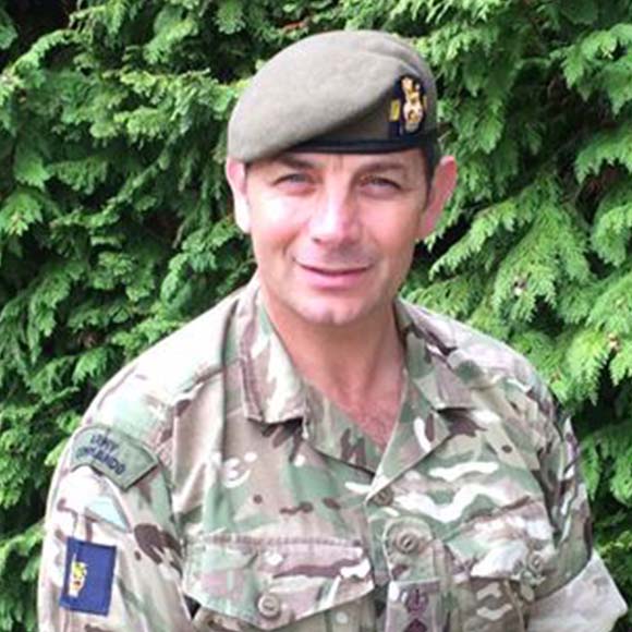 A man in military uniform and beret smiling, with a backdrop of green foliage.