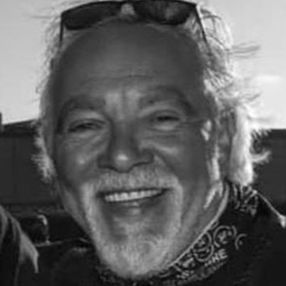 Black and white portrait of a smiling man wearing sunglasses on his head and a patterned scarf.