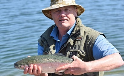 A man wearing a fishing hat, blue shirt, and green fishing vest holds a large rainbow trout near the edge of a calm lake. The sunny weather highlights the fish's shimmering scales and the serene water in the background.