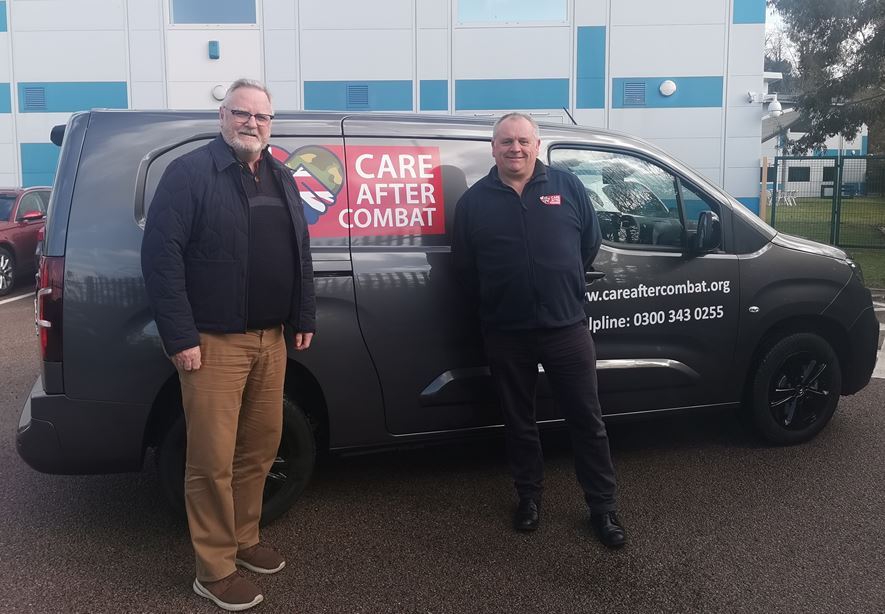 Two men standing in front of a grey van with 'Care After Combat' branding, including a logo featuring a handshake combining the British flag and camouflage pattern. The side of the van displays the website www.careaftercombat.org and the helpline number 0300 343 0255. The background includes a modern building and a fence.