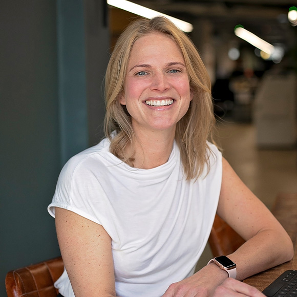 A smiling woman with shoulder-length blonde hair, wearing a white T-shirt and a smartwatch, seated indoors in a modern office setting.