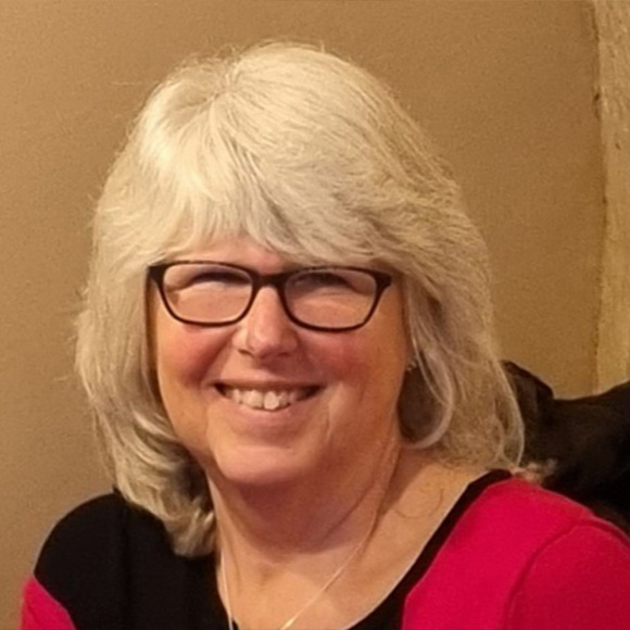 A smiling woman with silver hair wearing glasses, a red and black top, seated indoors against a beige wall.