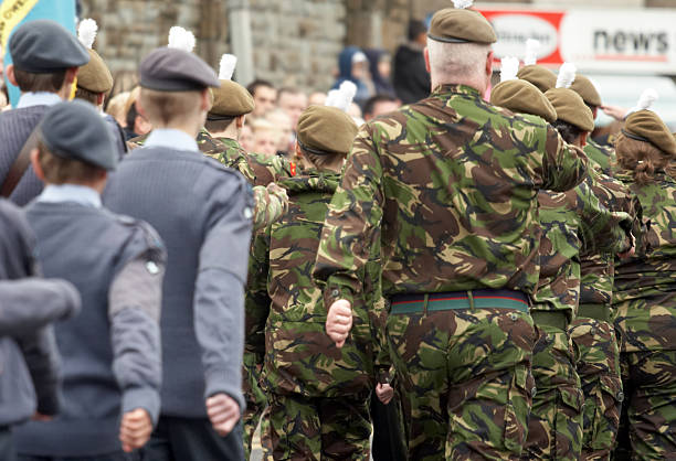 A group of uniformed military personnel and cadets marching in formation during a public parade, with people in camouflage and others in blue uniforms, set against a backdrop of a stone building and crowd.