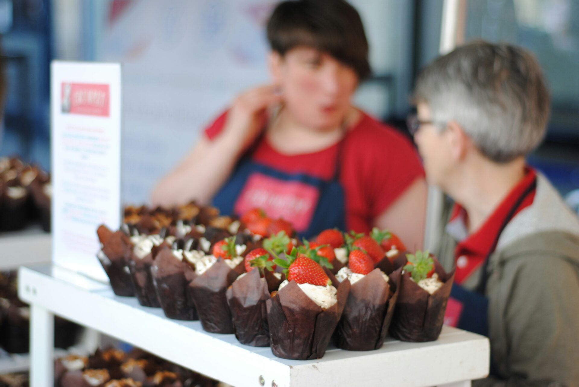 Close-up of a display of cupcakes topped with fresh strawberries, with two people in conversation in the blurred background wearing aprons.