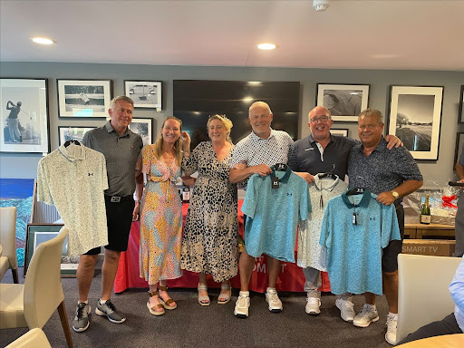 "A group of six people smiling and posing together indoors, holding up golf shirts. They are standing in front of a table displaying prizes, with framed pictures hanging on the wall behind them.