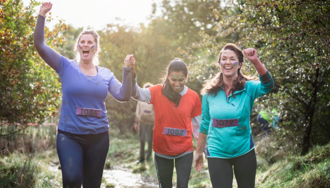 Three women smiling and celebrating as they finish an outdoor obstacle race, covered in mud and wearing race bibs, with a wooded area in the background.