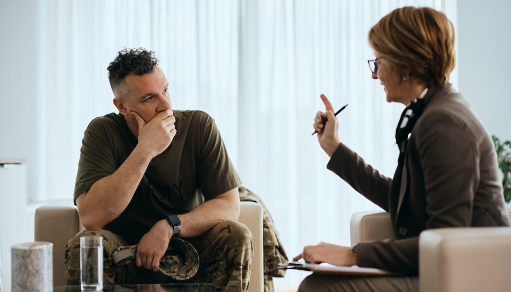 A military veteran in camouflage trousers and a t-shirt sits thoughtfully in a counselling session, listening to a professional wearing glasses and holding a pen, set in a well-lit office.