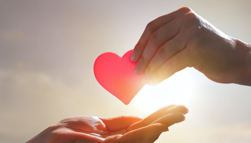 A bright red heart being passed from one hand to another, with sunlight shining in the background, symbolising giving and compassion.