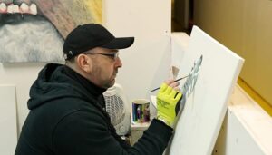 An artist wearing a black cap and glasses carefully painting on a canvas in a studio, with art supplies and a heater in the background.