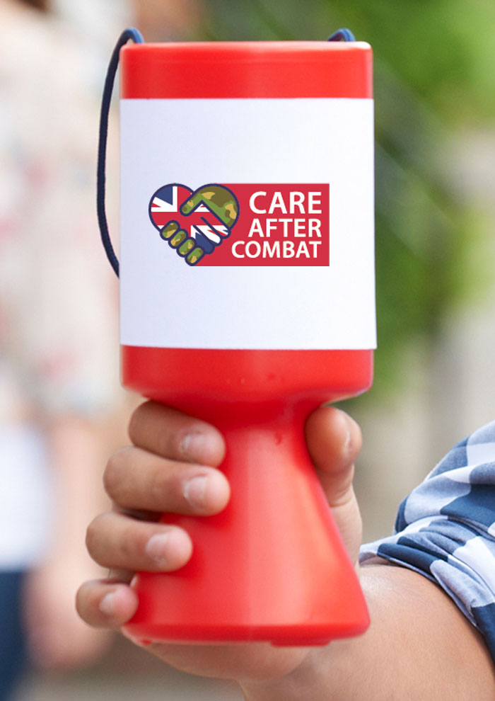 A close-up of a hand holding a red charity donation bucket labeled with the 'Care After Combat' logo, featuring a camouflage and Union Jack heart design, against a blurred outdoor background.