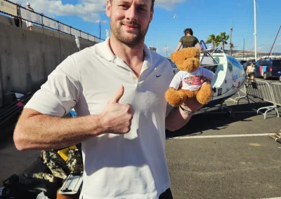 A man standing beside a small aircraft gives a thumbs-up while holding a teddy bear wearing a T-shirt, representing the Care After Combat initiative and the Operation PAWSIBLE campaign, with a marina and blue sky in the background.