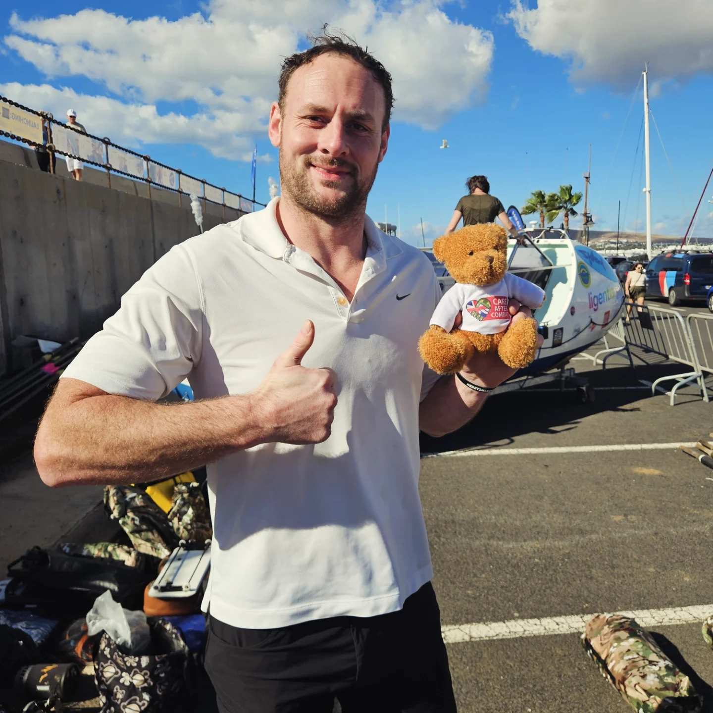 “A man standing beside a small aircraft gives a thumbs-up while holding a teddy bear wearing a T-shirt, representing the Care After Combat initiative and the Operation PAWSIBLE campaign, with a marina and blue sky in the background.