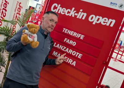 A man at a Jet2 check-in area holding a teddy bear wearing a Care After Combat T-shirt as part of the Operation PAWSIBLE campaign, pointing at a departures board showing holiday destinations, with luggage beside him in the airport.