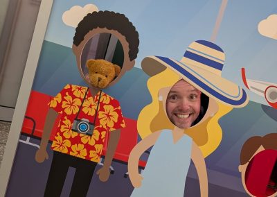 A man posing with a teddy bear wearing a Care After Combat T-shirt for the Operation PAWSIBLE campaign, placing their faces through a Jet2 holiday photo board at the airport, designed as a fun travel scene with cartoon passengers and suitcases.