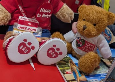 Two teddy bears sitting on an airport counter, one wearing a Jet2.com ‘Happy to Help’ uniform and the other wearing a Care After Combat T-shirt as part of the Operation PAWSIBLE campaign, with an airport desk and staff visible in the background.