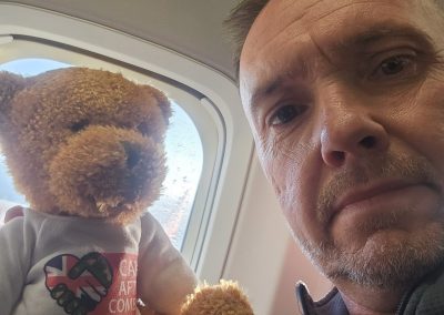 A man sitting on an aeroplane next to the window holding a teddy bear wearing a Care After Combat T-shirt, supporting the Operation PAWSIBLE campaign while travelling, with the aircraft window visible in the background.