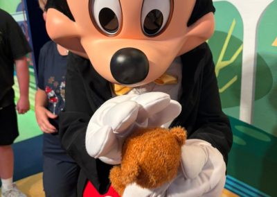 A person dressed as Mickey Mouse holding a teddy bear wearing a Care After Combat T-shirt for the Operation PAWSIBLE campaign, during a character meet-and-greet at a theme park.