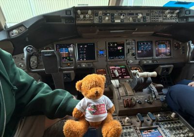Teddy bear wearing a Care After Combat T-shirt for the Operation PAWSIBLE campaign sitting inside an aeroplane cockpit surrounded by flight controls and instruments, with crew members seated on either side.