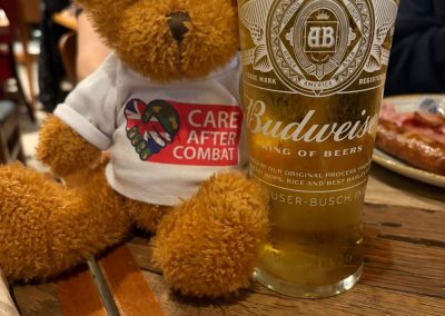 Teddy bear wearing a Care After Combat T-shirt as part of the Operation PAWSIBLE campaign sitting on a pub table next to a pint of beer, with food and a restaurant setting in the background.