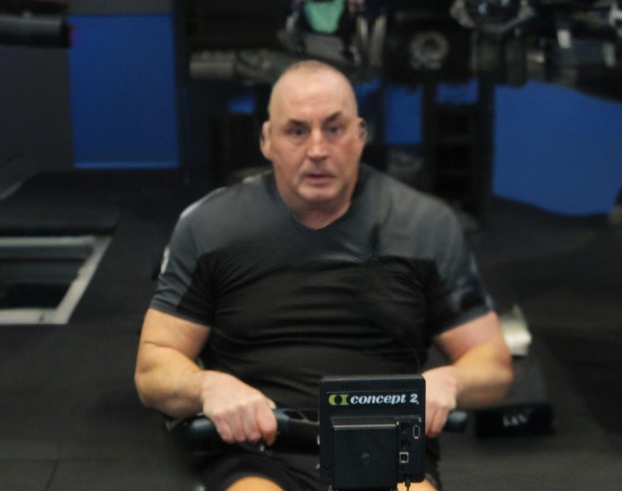 Man using an indoor rowing machine during a fitness training session, wearing a black sports shirt in a gym environment.