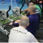 Group of veterans using indoor rowing machines during a training session in a gym, with participants wearing sports clothing, including a shirt labelled ‘Veteran’.