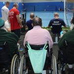 “Participants, including wheelchair users, taking part in an indoor rowing training session in a sports hall, with veterans and instructors gathered around rowing machines.