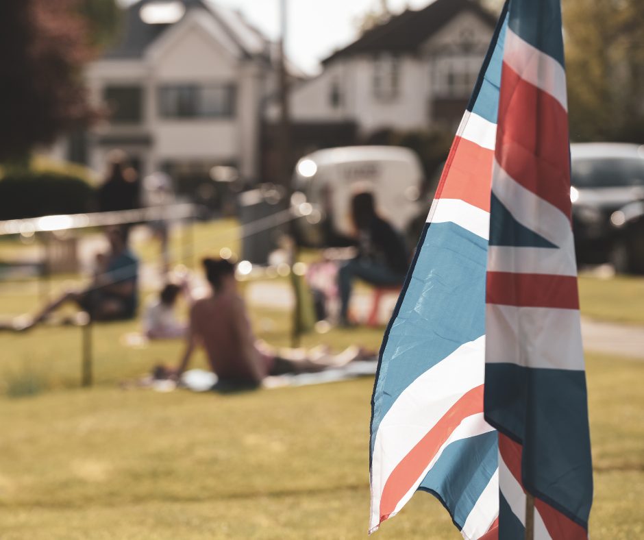 Union Jack flag in the foreground with people gathered outdoors in the background during a VE Day community event.